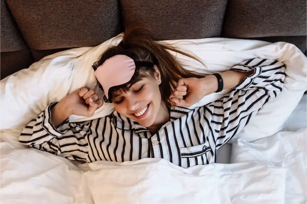 Girl in pajama and sleep mask is stretching and smiling while lying in bed